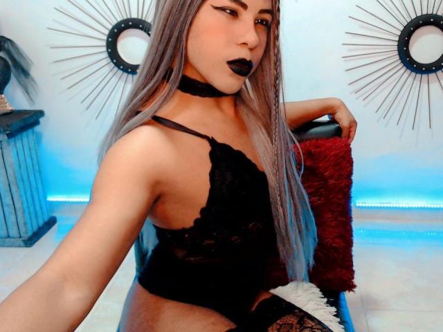 SakuraRiver, avec ses longs cheveux gris, sa lingerie en dentelle noire, son collier noir et son rouge à lèvres foncé, tenant un oreiller rouge, montrant ses seins, et un accessoire blanc et duveteux sur son genou..