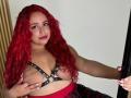 Estefy69 with red curly hair, black leather harness, holding her large tits, red lipstick, pink nails, smiling, standing by a door, wearing a red and black plaid skirt.