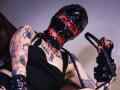 AmySpider in black latex mask with red laces, holding black leather flogger, wearing black tank top, showing tattoos, sitting on brown leather couch, exposing armpit hair.