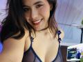 LucieMilner with brown hair, smiling, wearing a sheer blue top showing her tits, a black choker, and a floral strap. She's holding a phone for a selfie.