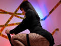 SkyeFrost with black dress, fishnet stockings, and yellow "Peligro" tape, squatting, showing her ass, in a dimly lit room with pink and blue lighting.