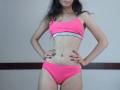 VikkiRi stands with hands on hips, wearing a pink sports bra and matching pink panties, showing a slim, flat stomach and small, firm tits. She has long, dark hair.