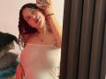 ValeryScot with curly red hair, wearing a sheer white top, showing her tits and nipples, taking a mirror selfie, smiling with red lipstick, wrist bracelet, and necklace.