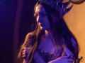 NikaFlorence with long brown hair, wearing a metal crown with pointy horns, and a jeweled, strapless top, gazing to the side, her hand on her chest, under blue-purple light.