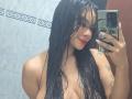 Yurimy with wet black hair, red lipstick, and tattoos, holding a phone in a mirror selfie, showing her bare, large tits. She's smiling, with a sexy look.