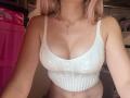 EliseMonet with pink hair, wearing a white ribbed crop top, showing her large tits and fair skin, a delicate necklace, and her arms slightly out.