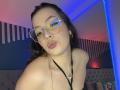VanessaClose with pouty lips, wearing glasses, hoop earrings, black necklace, and a halter top, showing her bare shoulders, with wavy brown hair and blue accent lighting.