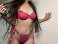 Tisia with long brown hair, wearing red lace bra and panty set with pink floral embellishments, holding hair, showing off her tits and pussy. Her hand touches a white wall.