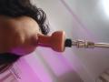 Chili's face with long dark hair, mouth around a pink dildo attached to a silver and black sex toy, visible nipple on the left.