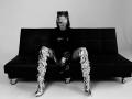 MilagrosEdmisten, a dark-skinned woman with braided hair and a mustache, sits on a black couch, wearing a black top and eye mask, and ornate patterned boots. Fucking sexy. High-contrast black-and-white photo.
