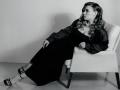 Niferttiti with wavy hair, wearing a black dress and shiny jacket, sits on a white chair, legs crossed, showing her tits, in black-and-white, looking to the side.