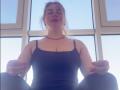 AvaQueen with her tits visible in a black tank top, sitting with legs apart, hands on knees, red necklace, and red nails, against a bright window.