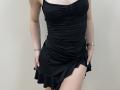 Ramilla's black ruched dress, showing her tits, with hands in pockets, wearing a gold bracelet, standing against a plain wall, legs slightly apart.