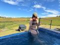 LaraHarvey with red hair, wearing a green cap and black thong bikini, sits in a blue-tiled pool, showing her butt, with a barbecue grill in the background.