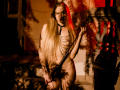 Naked MiaMeowa with long blonde hair, wearing chains over tits, holding a black strap, standing in dim red and yellow light, shadow patterns on her body.