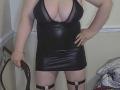 TitanicTits with big tits in a black, deep-v, leather mini dress, holding a whip, wearing black thigh straps, standing, hand on hip, in a dimly lit room.