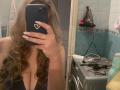 CharlineGusty with wavy brown hair, holding a phone covering her face, showing her cleavage, and a mirror selfie in a bathroom with a washing machine. Boobs visible.