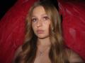 A photo of AkilahGhil with long blonde hair, pink eyeshadow, and a silver necklace, looking serious with red fabric in the background. She has small tits and a cute face.