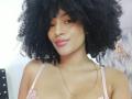 SnackTime has a big, fluffy afro, brown skin, a nose ring, pink lipstick, and a gold chain. She's wearing a pink bra with floral details.