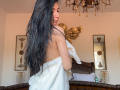 SinReea with long black hair, back to camera, wrapped in a white towel, holding a glove, standing in a room with a chandelier and decorative wall pieces.