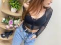 Rioria with long red hair, black sheer top, showing small boobs, and blue jeans, hands in pockets, black high heels on wooden shelf, white wall, flowers, green plant.