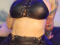 Kkole has big, shiny black leather boobs with lace-up details, showing off a tattooed arm and midriff, wearing matching black leather pants, under blue lighting.