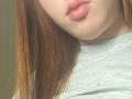 JannaCorsaro's red hair, pale skin, and pink lips are in the frame, wearing a gray shirt, with a subtle light source highlighting her upper chest.