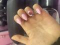 EliseMonet's hand with pink nails, one featuring a red gem and black pattern, against a black and pink background. Boobs are not visible.