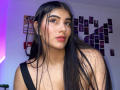 AlaraKey with long black hair, brown eyes, and red lipstick, wearing a black strap, showing her tits, standing in a room with photos on the wall.