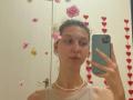 Sanella with flowers in her hair, wearing a beige top and pearl necklace, taking a mirror selfie with a green phone, showing a neutral face.