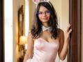 VikkiRi with blue glasses, pink satin bodysuit, white bunny ears, and fluffy tail, standing in a doorway, showing her tits and smiling. She has long dark hair.