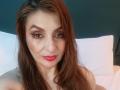ElenaHart has long brown hair, red lipstick, and brown eyes, with subtle makeup, and is looking at the camera with a slight smile, showing her bare shoulders.