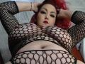 MirandaRossi with red hair, wearing a black fishnet top, showing huge tits and a belly button. Hands in hair, looking down. Red lipstick, sexy expression.