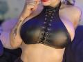 Kkole with big tits, wearing a black leather halter top with lace-up front, biting her red lipstick-covered lower lip. She has tattoos on her right arm.