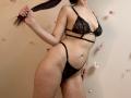 Tisia with black lace bra and thong, holding her long brown pigtails, showing her slim, toned body, small tits, and shaved pussy, standing in a slightly provocative pose.