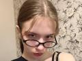 EviaMarcot with glasses, pale skin, brown hair in a ponytail, blue eyes, red lipstick, and a lip piercing, wearing a black top, looking directly at the camera.