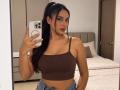 AdalinaVera with dark hair in a ponytail, brown ribbed crop top showing her tits, and denim pants, holding a phone in a mirror selfie, standing in a bedroom.