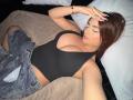 LaraHarvey with big tits, wearing a black crop top, lying on a bed, hand on head, denim shorts, brown hair, white nails, bracelet, pillow, and beige blanket.