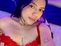 SophiaMarx with dark hair, light skin, and red lace bra, showing small tits, a chest tattoo, and a circular arm tattoo, smiling at the camera.