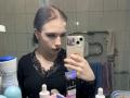 EviaMarcot with wet brown hair, dark lipstick, black lace top, holding phone, mirror selfie, bathroom counter with toiletries, looking at reflection, phone case with pink flower.