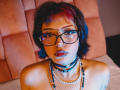 JadeMilleer with short black hair, red highlights, glasses, nose ring, lip piercing, choker, necklace, beads, and tattoos, looking at camera with blue and red light.