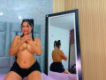 ExoticNativeLatina with dark hair in p Ponytails, topless, covering small tits with hands, wearing black shorts, reflected in mirror, standing by bed with purple sheets, wood wall.