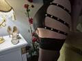 Rouzi trägt schwarze, trägerlose Dessous mit silbernen Nieten, die ihr Gesäß zeigen, und schwarze, spitzenbesetzte Overknees. Ein Paar beige High Heels steht auf einem weißen Tisch. Gedämpftes Licht..