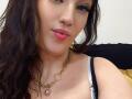 VanessaSquirtt with pink lips, brown wavy hair, black eyeliner, gold chain necklace, white strap, showing cleavage, smiling softly.