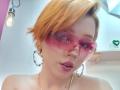 ChloeeWilliams with orange short hair, pink glasses, pink lipstick, and heart-shaped earrings, looking at the camera, showing her bare shoulders.