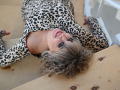 CandyLadyZ is lying on a wooden floor, wearing a leopard-print dress, with curly blonde hair, red lipstick, and a sexy smile, looking up at the camera.