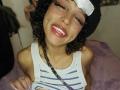 LunaKeller with curly black hair, wearing a black beanie, white striped tank top, and nose ring, smiling with red lipstick, blue makeup on forehead, lifting shirt to show tits.