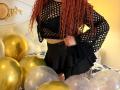 Niferttiti with curly red hair, black fishnet top, black ruffle skirt, and tattoo on thigh, standing among gold and silver balloons. Boobs visible through fishnet.