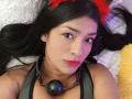ArianithaXox with long black hair, red furry cat ears, black ball gag, and chain, lying on a white fluffy pillow, wearing a grey top, with pink lipstick.