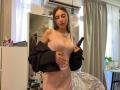 ErinMarrujo trägt ein pinkes Kleid, eine schwarze Jacke, die sie abgelegt hat, und hält ein Telefon. Sie trägt eine Halskette, hat braunes Haar und steht in einem Raum. Sie schaut nach unten. Im Hintergrund sind weiße Vorhänge zu sehen, und es gibt einen runden Tisch mit einer grünen Pflanze..